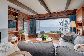 Un salón acogedor con un amplio ventanal con vistas al mar, vigas de madera en el techo, una chimenea y una estantería llena de libros.