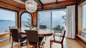 Comedor con mesa y sillas de madera, un candelabro y vista al mar a través de amplios ventanales.