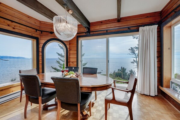 Comedor con mesa y sillas de madera, un candelabro y vista al mar a través de amplios ventanales.
