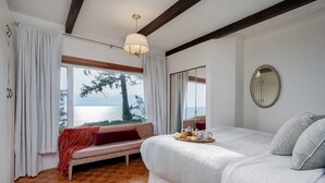 Un dormitorio con una cama grande, un banco con un cojín rojo, una bandeja con alimentos para el desayuno y vistas al mar.
