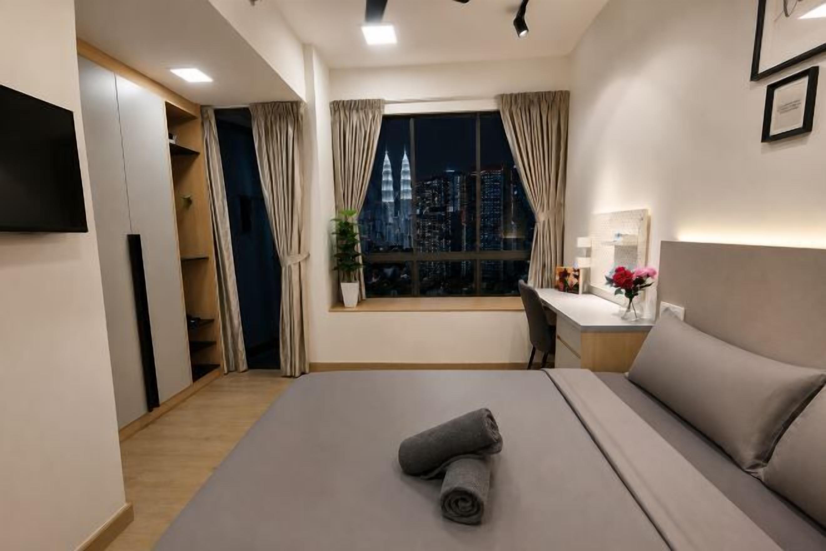 Et hotelværelse med seng, skrivebord, stol, fjernsyn, et vindue med gardiner og udsigt over byen.