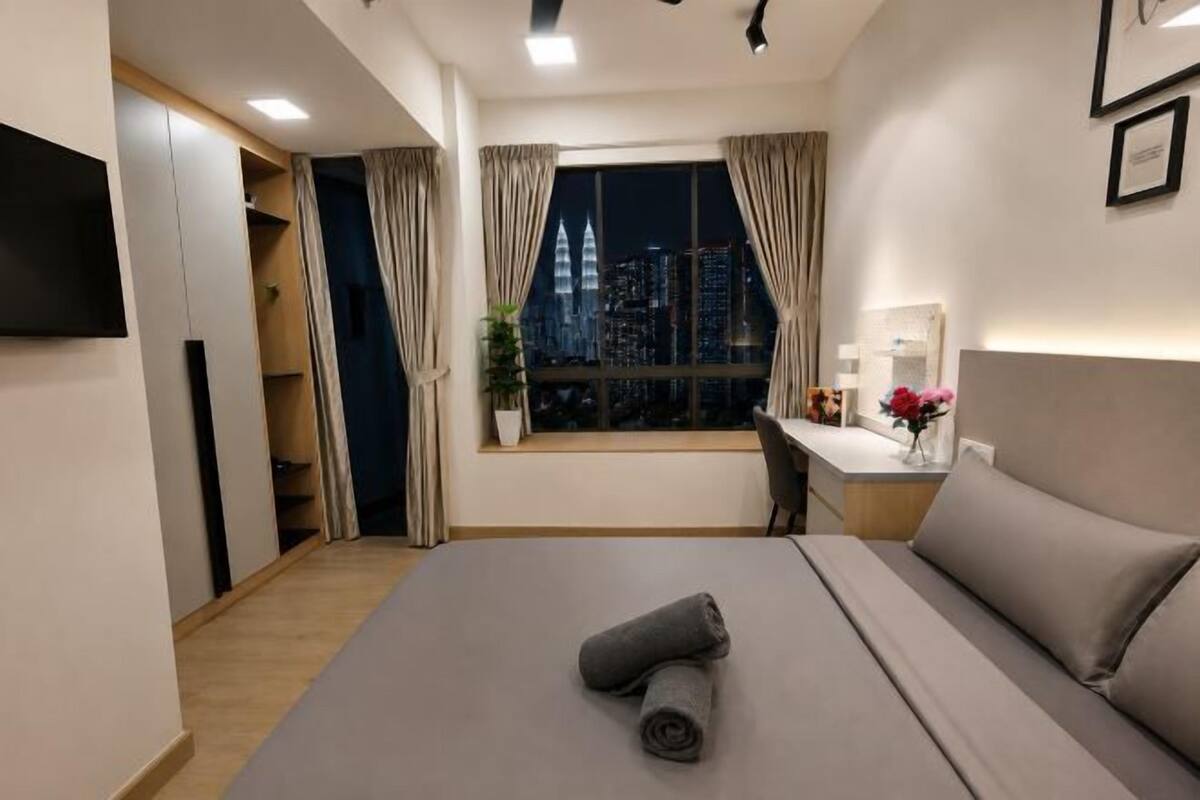 Habitación de hotel con cama, escritorio, silla, televisor, ventana con cortinas y vista a la ciudad.