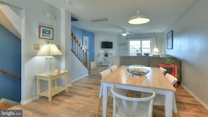 A dining area with a wooden table and white chairs, a staircase, and a living room with a sofa and a TV. - Spacious, family friendly 4-bedroom condo with pool in charming Dewey Beach!! (Dewey Beach)