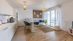 A modern kitchen with white cabinets, a dining area with blue chairs, and a large window with curtains. - Ty Enogat- Proche de la plage, garage privé (Dinard)