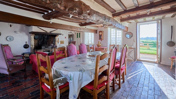 Een eethoek met een grote houten tafel, rode fauteuils en een rustiek plafond.