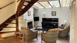 A living room with a wooden staircase, a fireplace, a piano, and two armchairs.