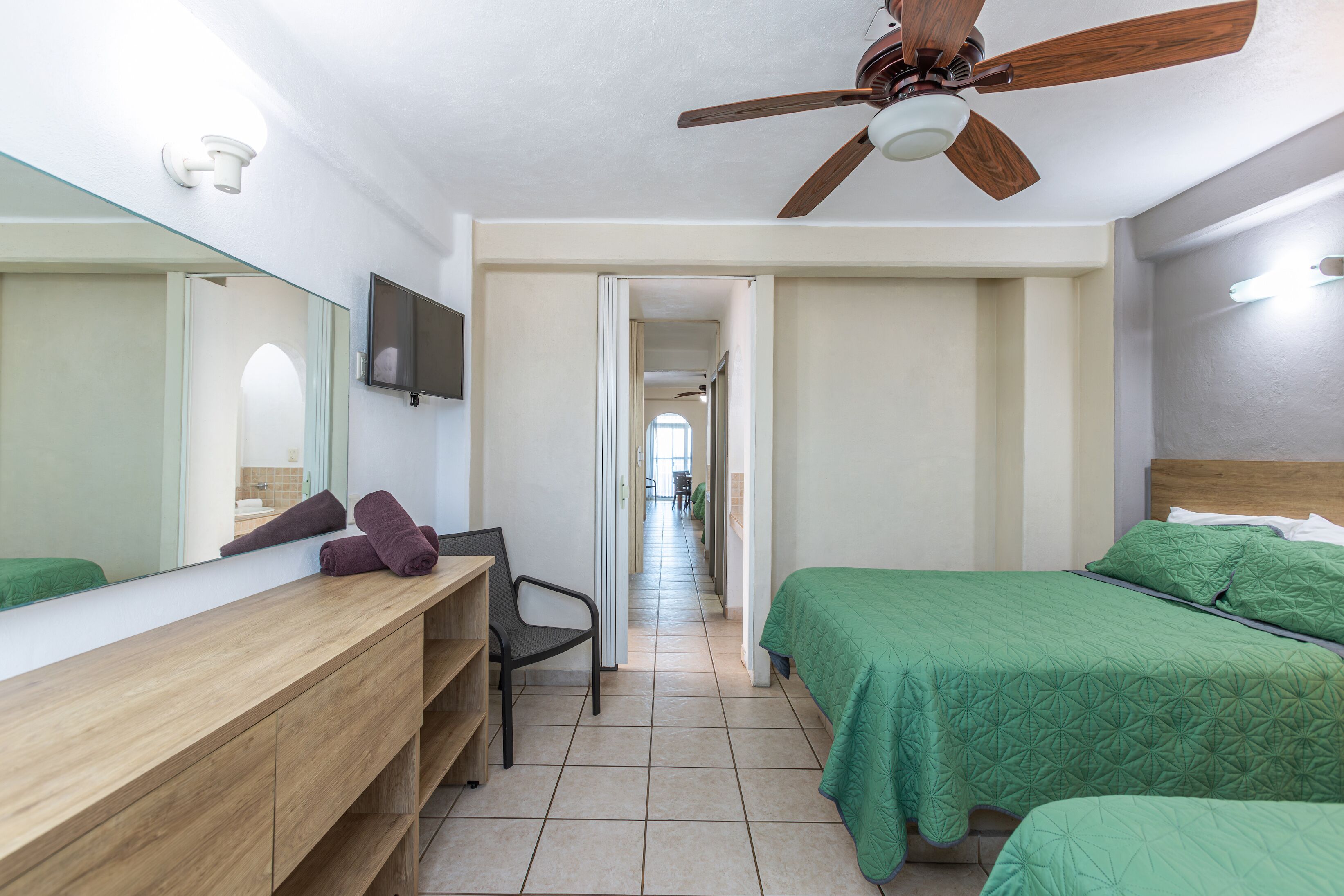 Un dormitorio con tocador de madera, cama verde, ventilador de techo y pasillo que conduce a otra habitación.
