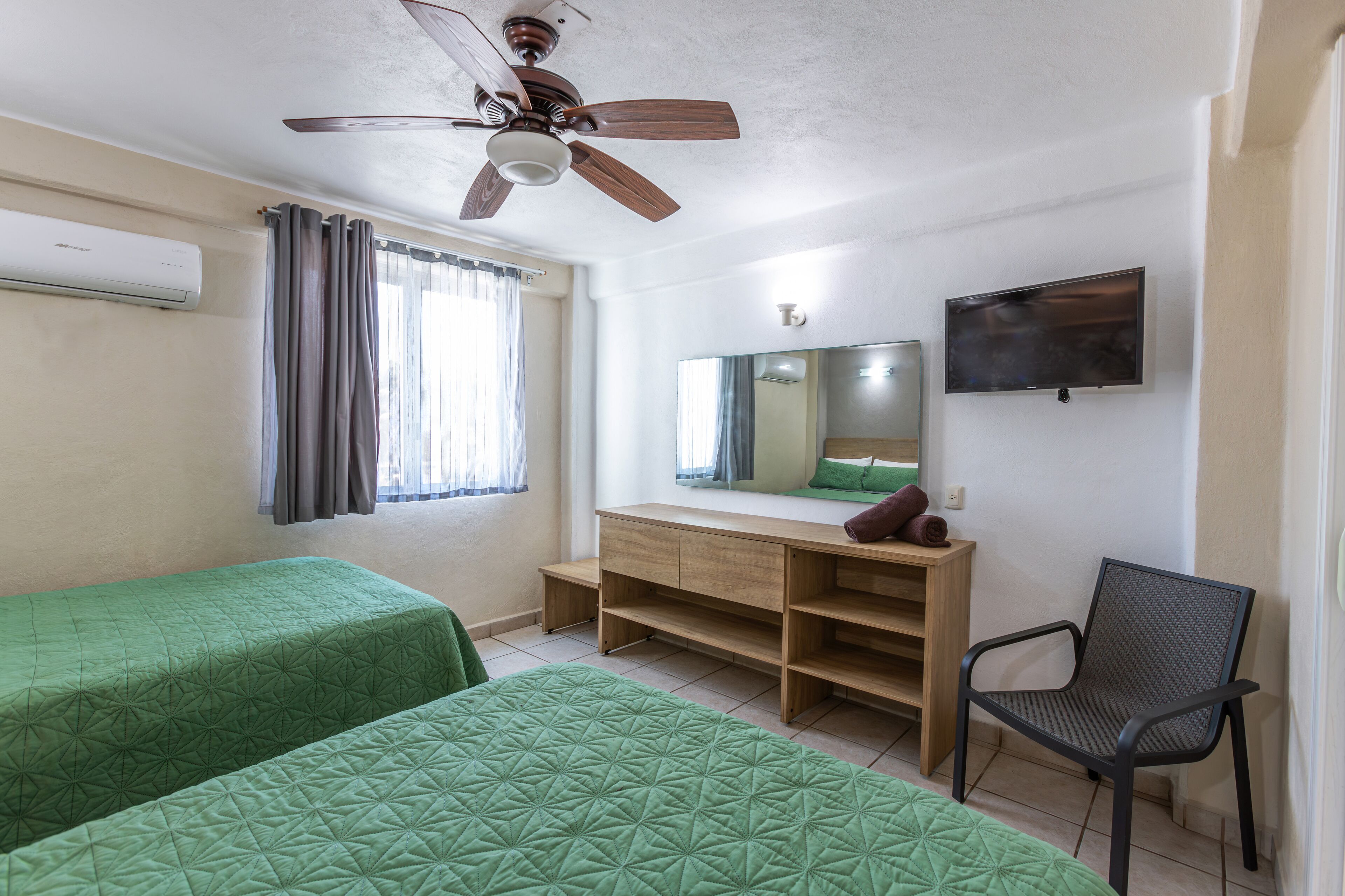 Habitación con dos camas, un escritorio, una silla, ventilador de techo, televisor y ventana con cortinas.