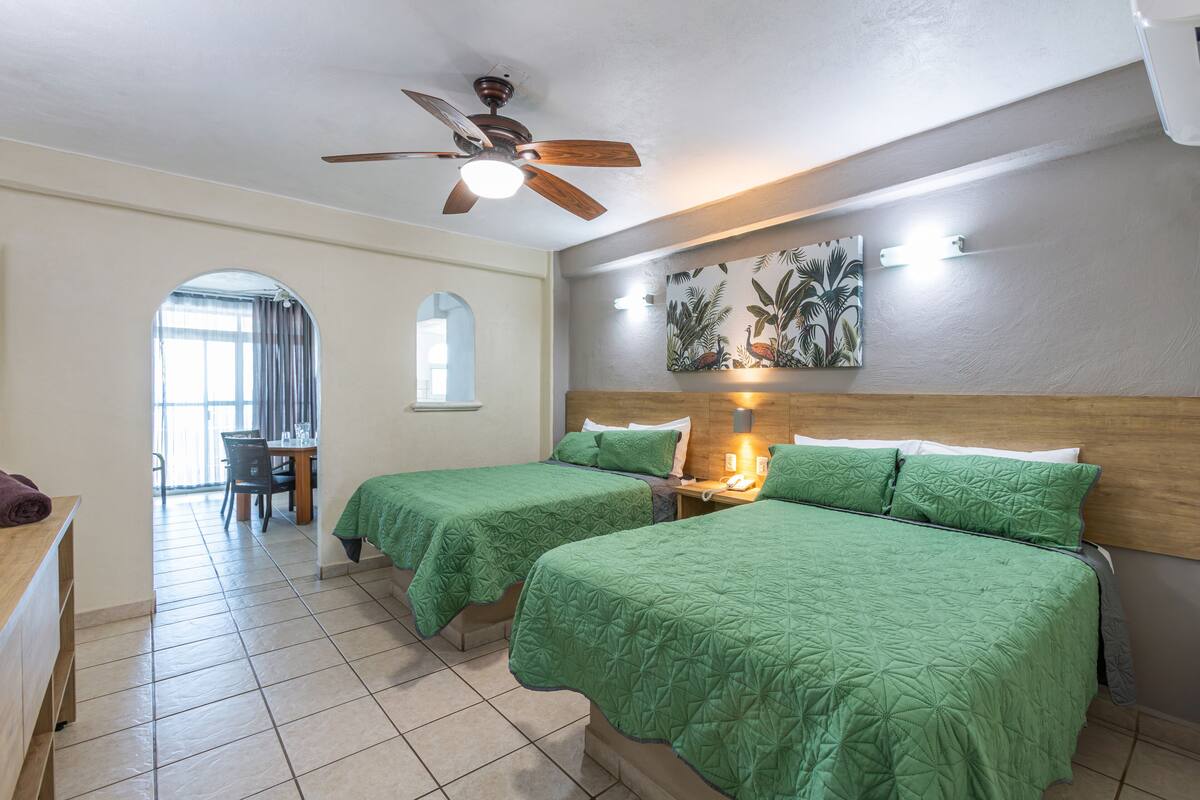 A hotel room with two beds, a ceiling fan, a dining area, and a view of the outdoors.