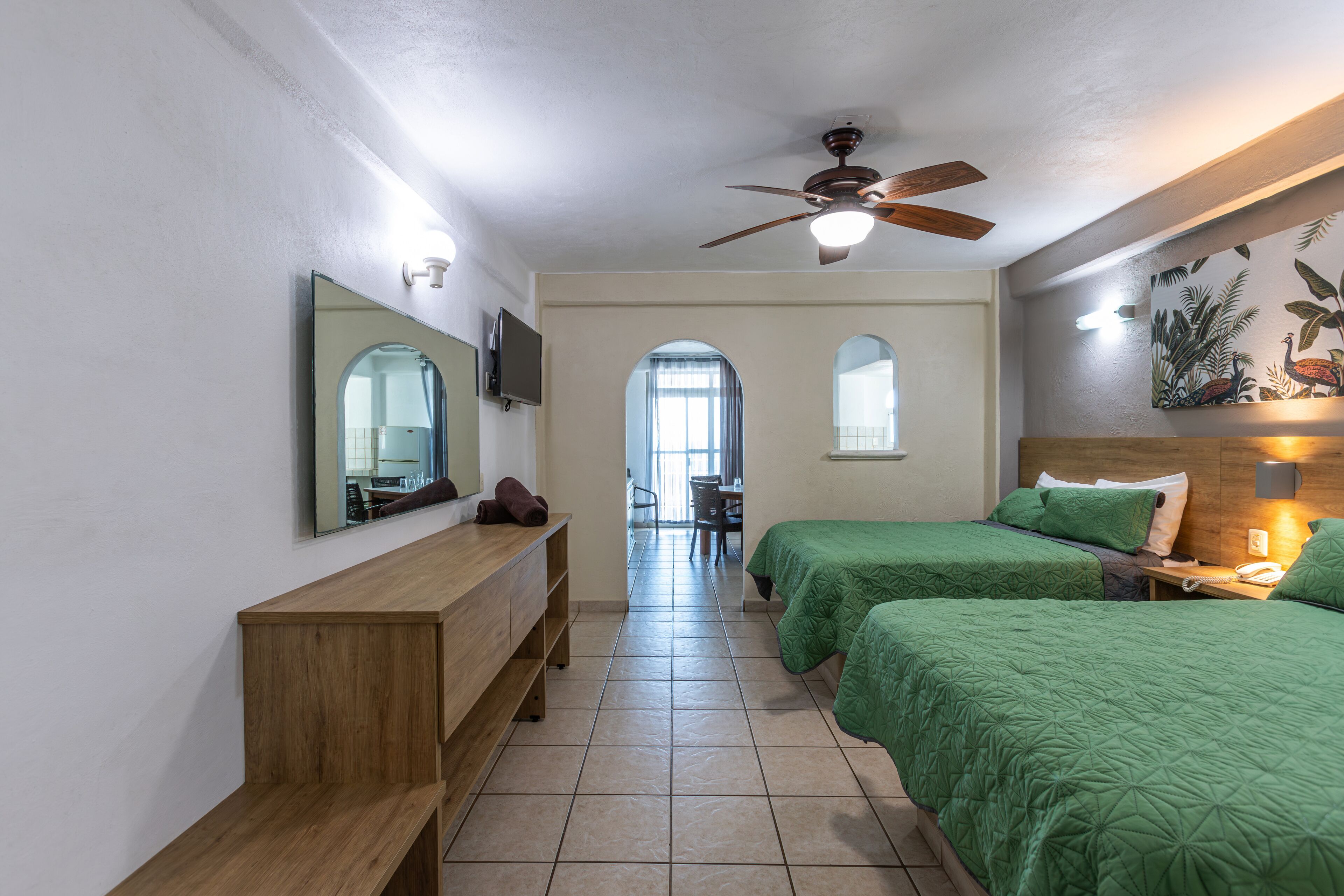 A hotel room with two beds, a wooden bench, a ceiling fan, a TV, and a painting on the wall.