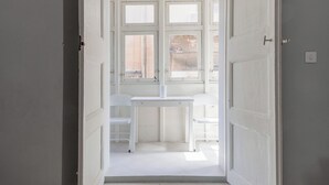 Une porte ouverte donne sur une pièce où se trouvent un bureau et une chaise blancs, une fenêtre et un sol carrelé.