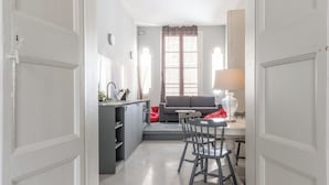 Un couloir mène à une salle à manger équipée d’une table et de chaises, d’un canapé et d’une fenêtre avec des rideaux.