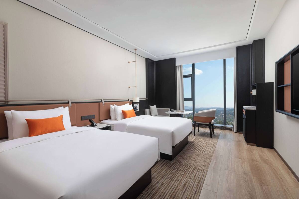 A hotel room with two beds, a desk, a chair, a TV, and a large window offering a city view.
