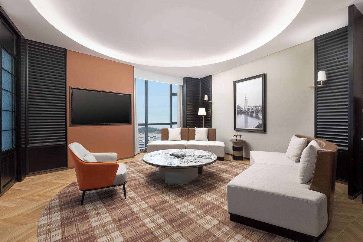 A modern hotel room with a large TV, two sofas, a coffee table, and a city view.