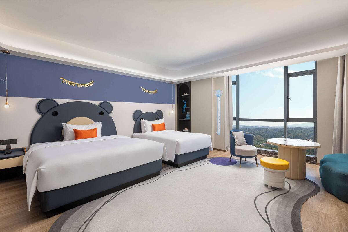 A hotel room with two beds, a desk, a chair, a small table, and a view of the outdoors.
