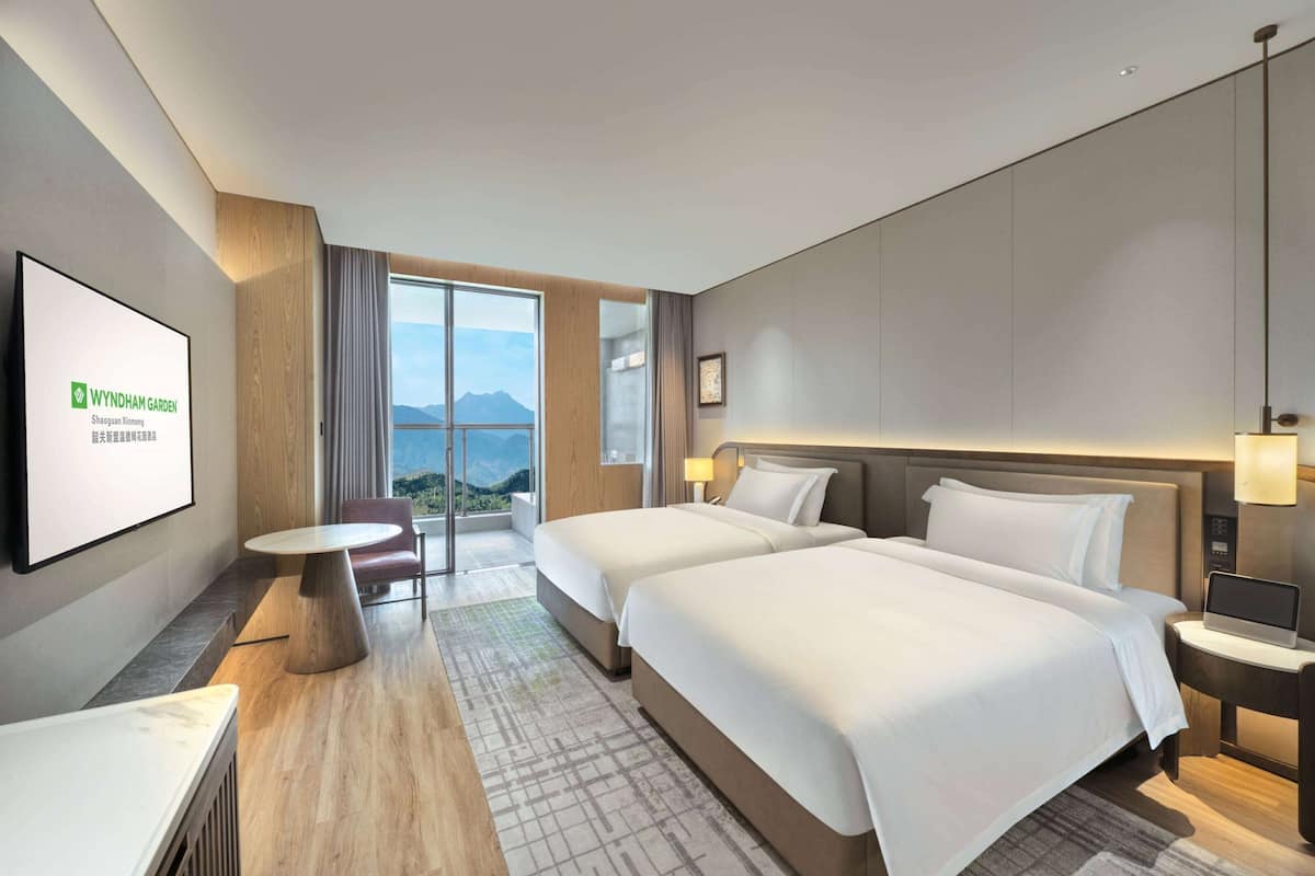 Een hotelkamer met twee bedden, een bureau, een stoel en een groot raam met uitzicht op de bergen.