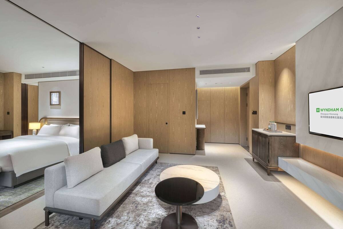 Een moderne hotelkamer met een bed, een bank, een ronde salontafel en een televisiescherm waarop 'WYNDHAM G' staat.