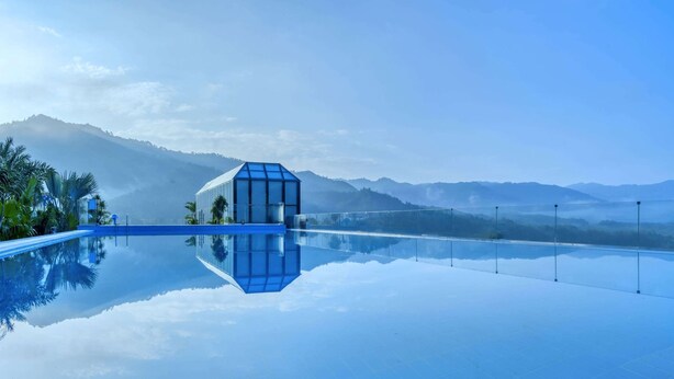 Une piscine d’un bleu éclatant, avec une structure moderne en verre au centre, entourée de palmiers et dominée par des montagnes en arrière-plan.