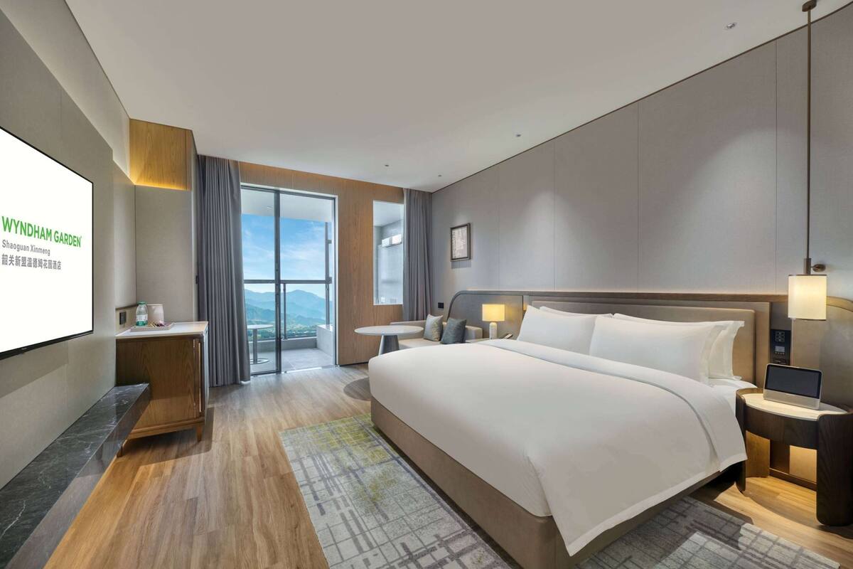 Een moderne hotelkamer met een groot bed, een bureau, een laptop en een televisiescherm waarop de naam van het hotel staat.