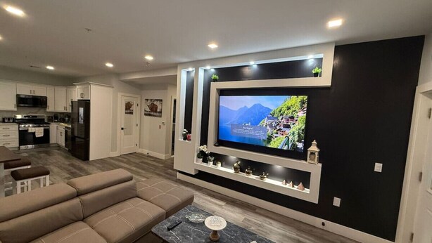 A modern living room with a large flat-screen TV, a sofa, a coffee table, and a kitchen in the background. - The Luxury Suite: 1BR Perfect for Long Stays (Paterson)