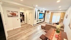 A modern living room with a sofa, a dining table with a red table runner, and a TV mounted on the wall. - The Luxury Suite: 1BR Perfect for Long Stays (Paterson)