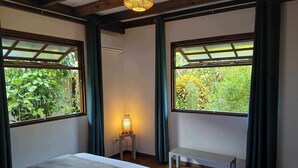 Une chambre à coucher avec un lit, une table de chevet avec une lampe, un banc et de grandes fenêtres avec des rideaux.