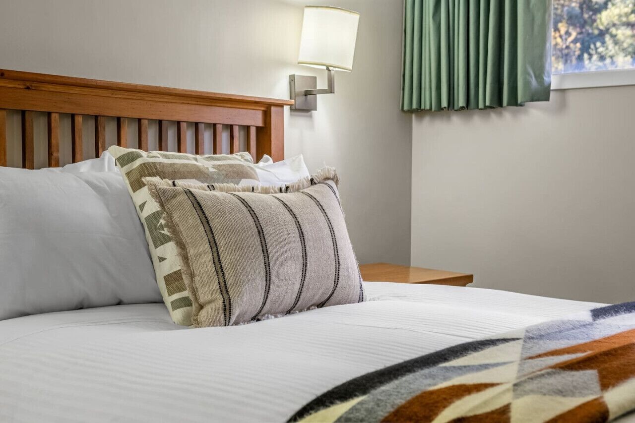 A neatly made bed with a striped pillow and a plain pillow, a wooden headboard, a bedside lamp, and a window with curtains.