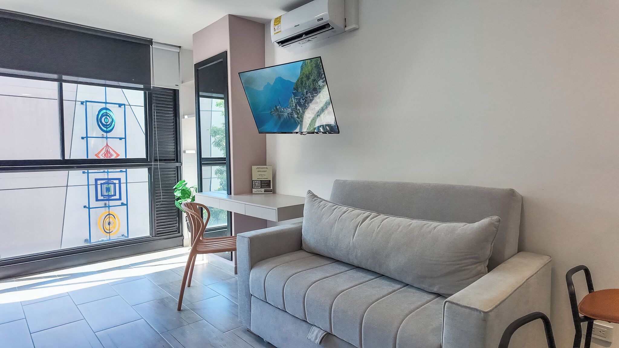 A modern living room with a grey sofa, a flat-screen TV mounted on the wall, a small table, and a chair.