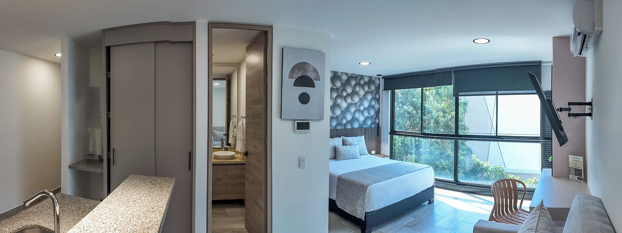 Ein modernes Schlafzimmer mit einem großen Bett, Blick auf Grün hinter einem Fenster und einem Badezimmer mit Dusche und Waschbecken.