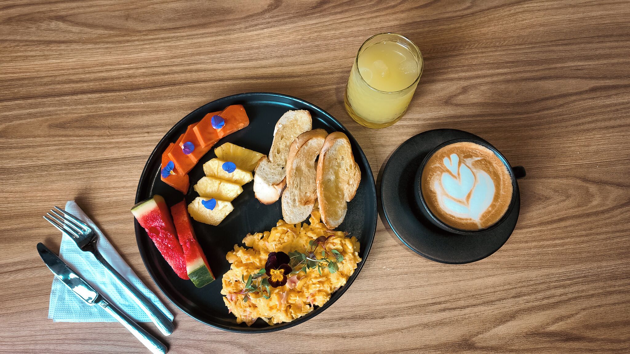 A breakfast plate with scrambled eggs, pineapple, watermelon, and toast, a glass of orange juice, and a cup of coffee with latte art on a wooden table.