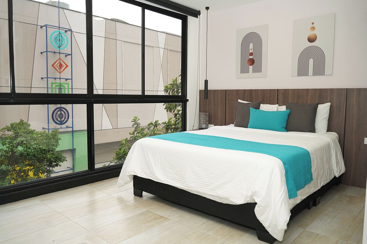 Een moderne slaapkamer met een groot raam, een bed met een wit en blauw beddegoed, en twee kunstwerken aan de muur.