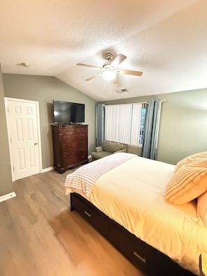 A bedroom with a bed, a dresser, a ceiling fan, a television, and a window with curtains.