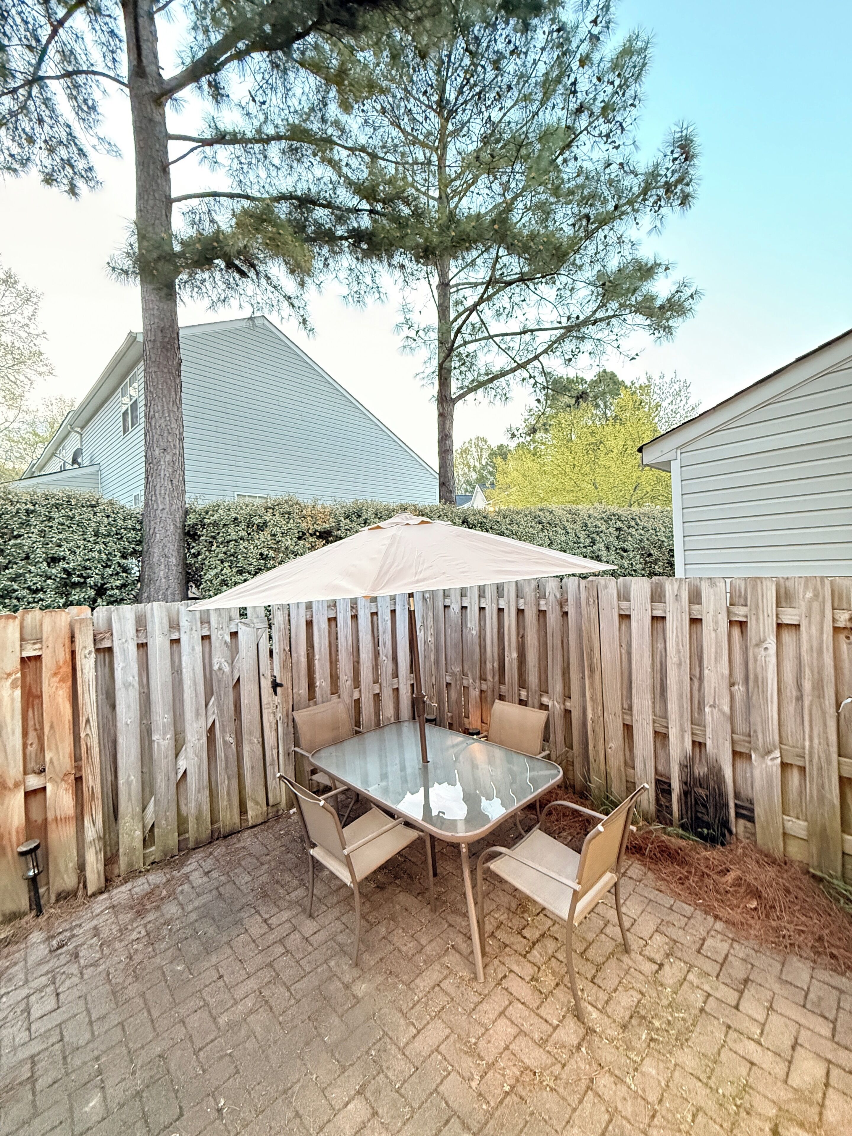 Une terrasse avec une table et des chaises en verre, une clôture en bois et une haie.