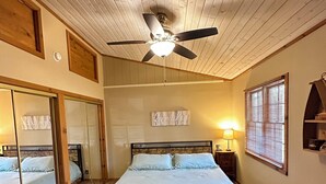 Et soveværelse med en seng, en ventilator i loftet, et natbord med lampe, et spejl og et vindue med persienner.