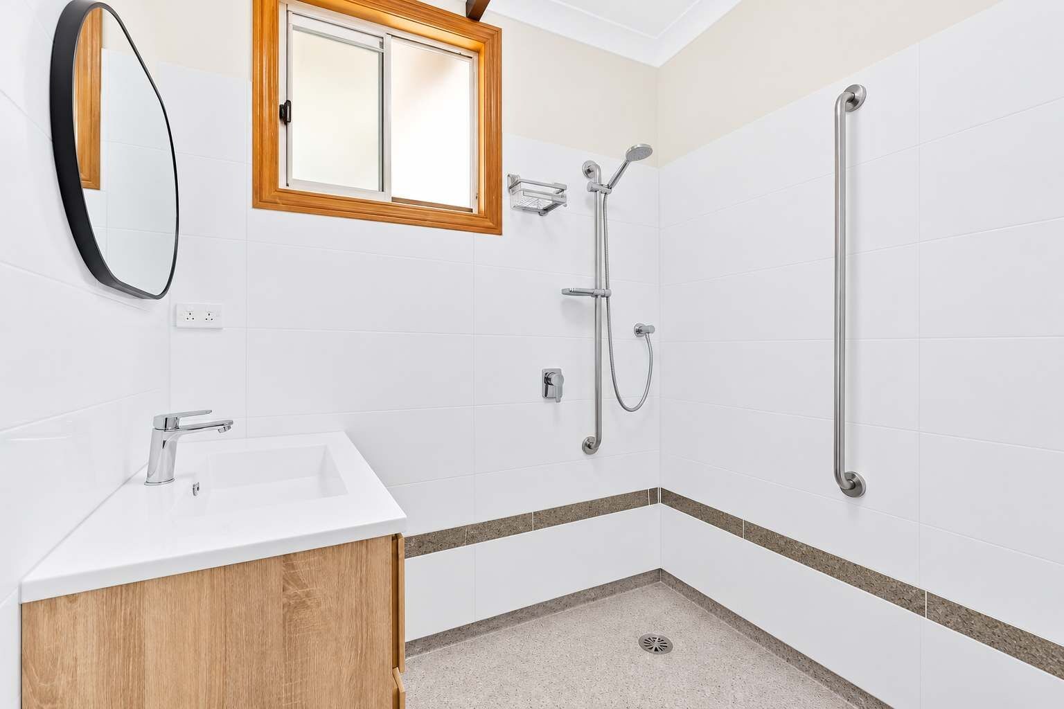 Une salle de bain moderne avec un lavabo blanc, un miroir, une douche équipée d’un pommeau de douche à main et d’une barre d’appui.