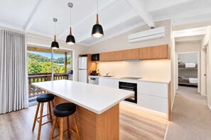 A modern kitchen with a central island, wooden stools, pendant lighting, and a view of greenery outside.