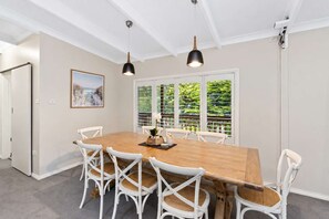 Un comedor con una mesa de madera y sillas blancas, dos lámparas colgantes, una ventana con persianas y un cuadro en la pared.