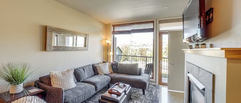 En moderne stue med en hjørnesofa, en pejs og et stort vindue med udsigt til en balkon.