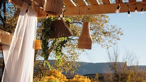Una pérgola con lámparas de mimbre colgantes y cortinas blancas, frente a un telón de fondo de follaje otoñal y colinas lejanas.