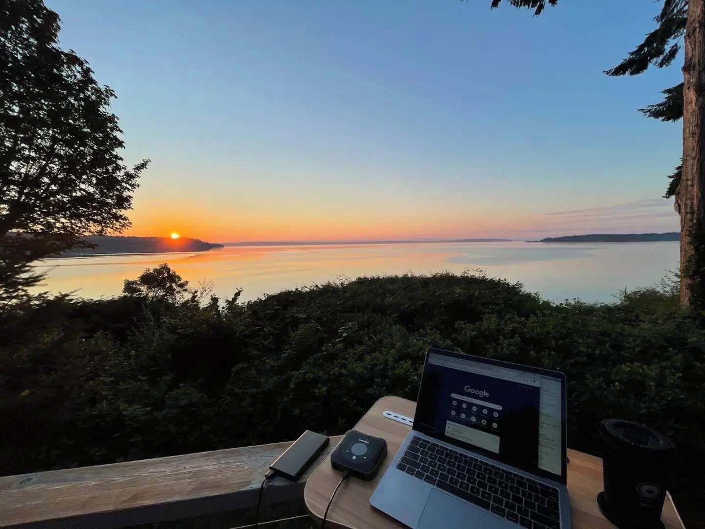 Um laptop sobre uma mesa de madeira, com vista para um pôr do sol sobre a água.