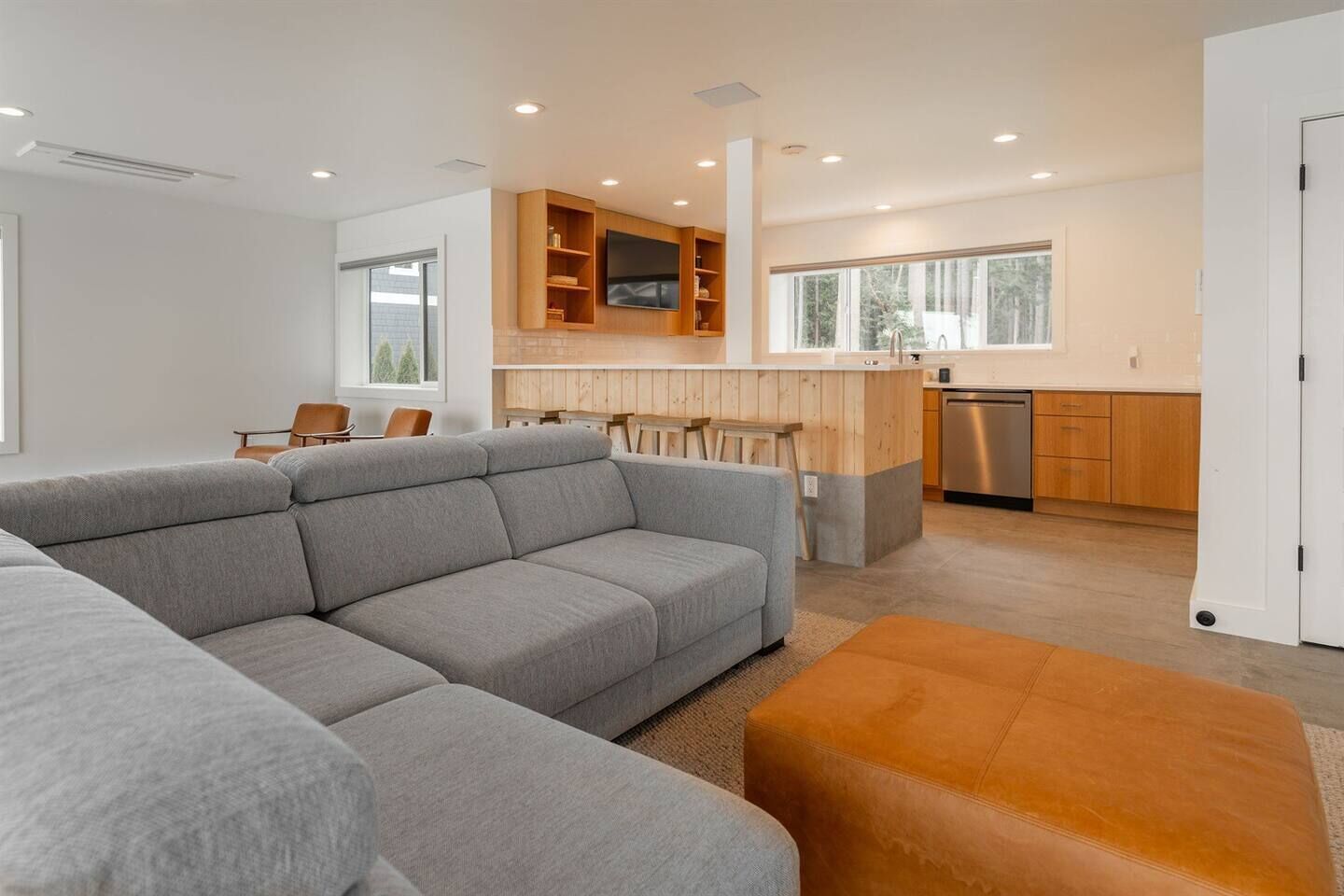 A modern living room with a grey sectional sofa, a wooden dining table, and a kitchen area with wooden cabinets and a built-in oven.
