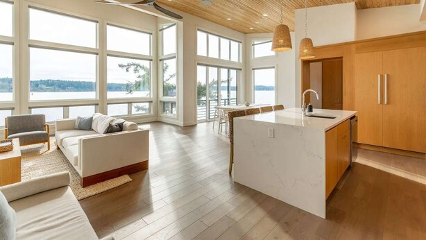 Een ruime woonkamer met een groot raam met uitzicht op een waterpartij, een houten plafond en een moderne keuken met houten keukenkastjes.