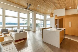 Een ruime woonkamer met een groot raam met uitzicht op een waterpartij, een houten plafond en een moderne keuken met houten keukenkastjes.