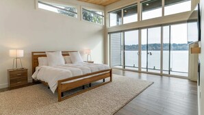 Een slaapkamer met een groot bed, een houten hoofdbord, twee nachtkastjes met lampen, een groot raam met uitzicht op een wateroppervlak, en een houten vloer.