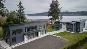Een modern huis met twee verdiepingen, een garage, grote ramen en uitzicht op een meer.