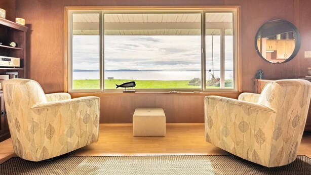 A room with two patterned armchairs facing a large window with a view of a grassy field and a body of water. - An Exciting Salish Escape w/ Waterfront View (Kingston)