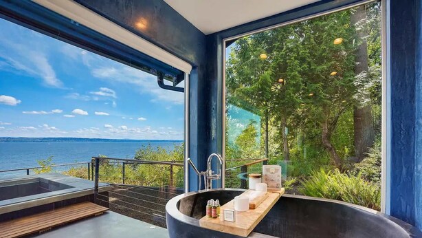 Een moderne badkamer met een groot zwart bad, houten terras en uitzicht op een meer en bos.
