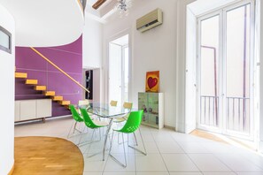 Uma área de refeições moderna com mesa de vidro e cadeiras verdes, parede de destaque roxa, escada e varanda com guarda-corrimão.
