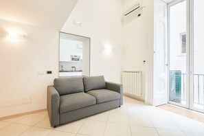A modern living room with a grey sofa, a large mirror, and a view of the kitchen and balcony.