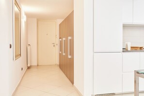 A hallway leading to a door, a kitchen area with white cabinets, and a small table.